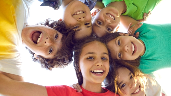 A group of smiling children all leaning over the camera in a circle with their hands on each others' shoulders