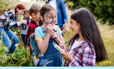 A group of children outdoors with grasses/plants on either side, the child nearest the camera is turned around smiling at her friend while holding a magnifying glass to her eye, the friend is taking notes on a paper notebook
